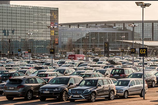 Voiture stationnée dans un parking longue durée à l'aéroport de Paris-Roissy-Charles-de-Gaulle en été, avec panneaux de terminaux et avion au décollage en arrière-plan